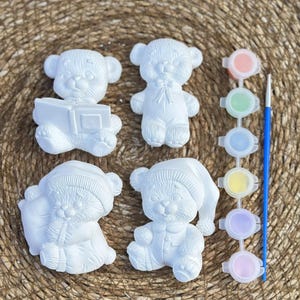 Op de afbeelding: Vier witte gipsen teddybeerbeeldjes in verschillende poses, klaar om te worden beschilderd. Eén beer houdt een boek vast, een ander draagt een nachtmuts en een derde heeft een pyjama aan. Verfpotten met diverse kleuren en een blauwe kwast zijn inbegrepen.