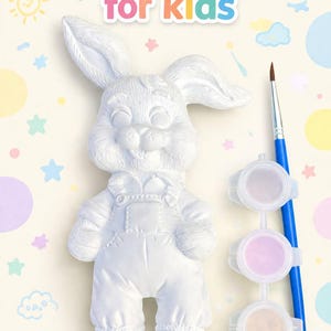 Può includere: Un kit di pittura per bambini con una figura di coniglio in gesso bianco con una salopette. Il kit include un pennello e tre vasetti di vernice dai colori pastello. Il testo "Painting kit for kids" è in alto.