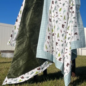 May include: A double-sided blanket featuring a solid dark olive green plush side and a white side with a repeating pattern of brown and light green dinosaurs, palm trees, and volcanoes, finished with a light blue plaid border trim.