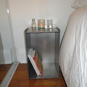 May include: A brushed metal bedside table. On top, there's a perfume bottle, a candle in a glass, and a decorative glass. Two books are inside the table. The table is approximately 40 cm wide.