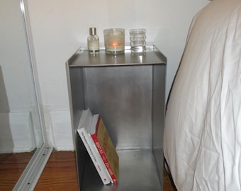 May include: A brushed metal bedside table with a rectangular shape. The top surface holds a small perfume bottle, a lit candle in a glass, and a decorative glass. Two books lean against the inside of the table.