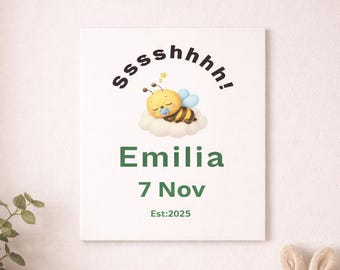 May include: A white canvas print with the name "Emilia" in green, along with the date "7 Nov" and "Est:2025". The print features a cartoon bee sleeping on a cloud. A wooden dresser holds a rainbow, star, and a basket with a stuffed bunny.