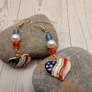 May include: A pair of patriotic heart-shaped earrings. The earrings feature a gold-tone hook, blue and red beads, and a heart pendant with a white, red, and blue design, embellished with rhinestones. The design resembles the American flag.