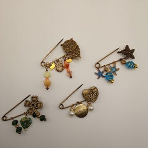 May include: Four antique bronze-toned safety pins with decorative charms. One pin features an owl, another a four-leaf clover, a third has starfish and blue beads, and the last has a heart with the word "Wish".