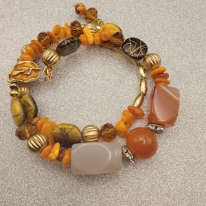 May include: A multi-strand wrap bracelet featuring a variety of beads in shades of orange, brown, and gold. The design includes smooth rectangular stones, ribbed gold-tone metal spheres, translucent amber-colored beads, and a gold-tone leaf charm.