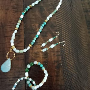 May include: A jewelry set featuring a necklace with a teardrop pendant, matching earrings, and two bracelets. The set includes turquoise, white, and green beads with gold accents. The jewelry is displayed on a dark wooden surface.