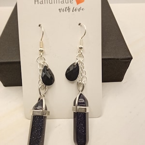 May include: A pair of dangle earrings with dark blue, glittery, crystal-like pendants. The earrings have silver-colored chains and hooks, with a small black teardrop-shaped bead above each pendant. The tag reads "Handmade with love."