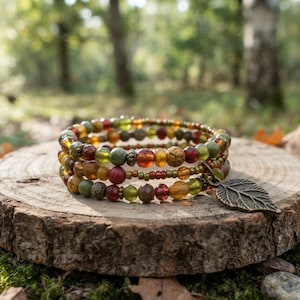 May include: A multi-strand wrap bracelet featuring a variety of round beads in shades of olive green, amber, deep red, and brown. The bracelet includes a metallic bronze-toned charm shaped like a leaf, attached to the end of the beaded strand.