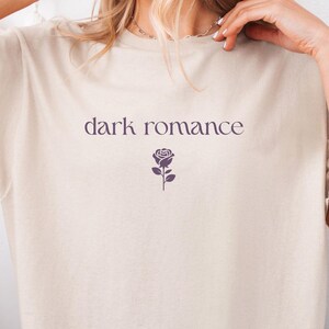 Könnte beinhalten: Beiges T-Shirt mit dem Aufdruck "dark romance" in dunklem Lila und einer Rosen-Grafik. Das Shirt hat eine lockere Passform und kurze Ärmel. Die Person trägt zerrissene blaue Jeans.