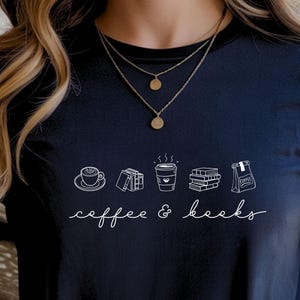 Könnte beinhalten: Marineblaues T-Shirt mit einer weißen Grafik von Kaffee und Büchern. Die Worte "coffee & books" sind in Kursivschrift unter den Illustrationen geschrieben. Das T-Shirt hat einen Rundhalsausschnitt und kurze Ärmel.