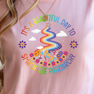 Könnte beinhalten: Hellrosa T-Shirt mit dem Text "It's a beautiful day to smash the patriarchy". Die Grafik zeigt einen Regenbogen, Wolken, Blumen und einen Stern. Das Shirt hat kurze Ärmel.