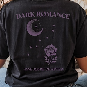 Könnte beinhalten: Schwarzes T-Shirt mit dem Schriftzug "DARK ROMANCE" über einem Mond und Sternen-Design. Darunter eine Rose und der Text "ONE MORE CHAPTER". Das Shirt hat einen Vintage-Look.