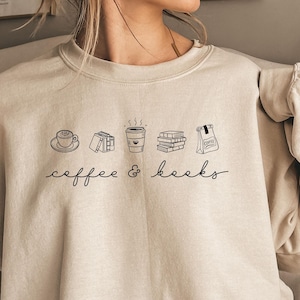 Könnte beinhalten: Ein beigefarbenes Sweatshirt mit Rundhalsausschnitt und einer schwarzen Strichzeichnung, die eine Kaffeetasse, Bücher und eine Kaffeetüte zeigt, mit dem Text "coffee & books" in einer kursiven Schriftart unter den Illustrationen.