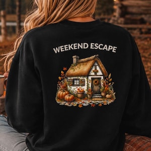 Könnte beinhalten: Ein schwarzes Langarm-Sweatshirt mit einem Motiv auf der Rückseite, das ein Häuschen mit Strohdach zeigt, umgeben von Kürbissen und Herbstblättern. Der Text "WEEKEND ESCAPE" ist in Weiß über der Illustration aufgedruckt.