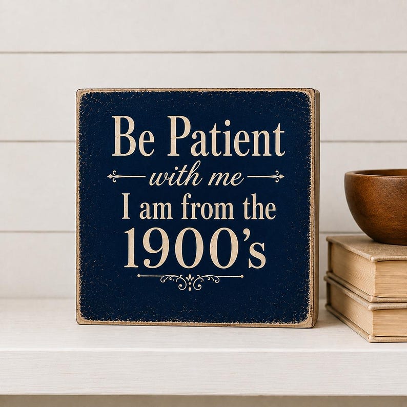 Dark navy wood shelf sitter with cream lettering reading “Be Patient With Me, I Am From the 1900s,” styled on a white shelf with neutral books and wooden bowl.