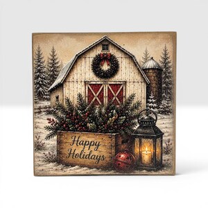 Puede incluir: Un letrero de madera cuadrado con una escena de granero invernal, una corona, una caja llena de ramas de pino y bayas, un cascabel rojo y un farol negro con una vela encendida. El texto Happy Holidays est&aacute; impreso en la parte frontal de la caja.