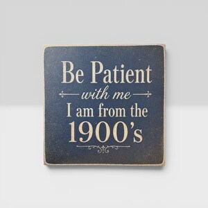 K&ouml;nnte beinhalten: Ein quadratisches, dunkelblaues Holzschild mit abgenutzten Kanten und dem wei&szlig;en Text Be Patient with me I am from the 1900's. Das Schild enth&auml;lt dekorative Verzierungen &uuml;ber und unter der mittleren Textzeile sowie ein Schn&ouml;rkelmuster am unteren Rand.