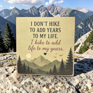 Könnte beinhalten: Ein quadratisches Holzschild mit hellbraunem Hintergrund und schwarzem sowie dunkelrotem Text: I DON'T HIKE TO ADD YEARS TO MY LIFE. I hike to add life to my years. Unter dem Text befindet sich eine Grafik mit grünen Bergen, Tannen, einer Sonne und Vögeln.