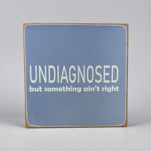 Könnte beinhalten: Quadratisches Holzschild mit hellblauem Hintergrund. Der weiße Text lautet "UNDIAGNOSED" über "but something ain't right". Das Schild hat einen braunen Rand, was auf einen Dekorationsartikel hindeutet.