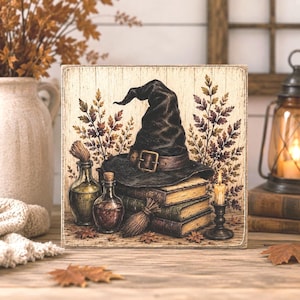 May include: A square wooden sign featuring a printed illustration of a black witch hat resting on a stack of three old books. Beside the books are two glass potion bottles, a small broom, and a lit candle in a dark metal holder, all framed by autumn leaves.