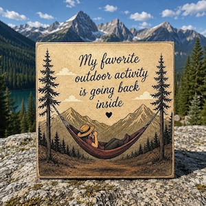 Könnte beinhalten: Ein quadratisches Holzschild mit einer aufgedruckten Illustration einer Person mit Hut, die in einer Hängematte zwischen zwei Kiefern liegt. Der Text lautet, My favorite outdoor activity is going back inside, mit einem kleinen schwarzen Herz.