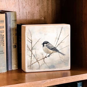 May include: A square wooden block featuring a painted illustration of a small bird with a black head and grey wings perched on thin, bare branches against a soft, muted, light-colored background, displayed next to vintage books.