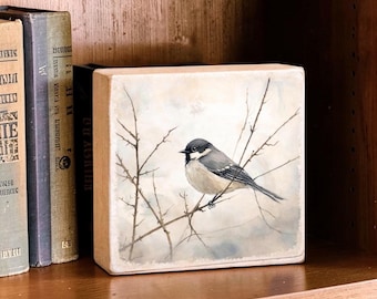 May include: A square wooden block featuring a painted illustration of a small bird with a black head and grey wings perched on thin, bare branches against a soft, muted, light-colored background, displayed next to vintage books.