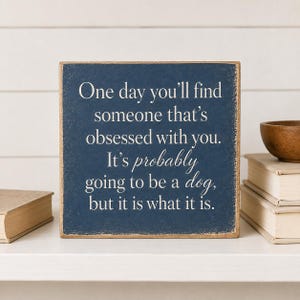 Könnte beinhalten: Ein quadratisches, marineblaues Holzschild mit abgenutzten Kanten und weißem Text: One day you'll find someone that's obsessed with you. It's probably going to be a dog, but it is what it is. Das Schild hat einen Rand aus natürlichem Holz.