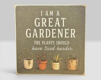 Może przedstawiać: Kwadratowy drewniany znak z ciemnoszarym tłem i białym napisem I AM A GREAT GARDENER THE PLANTS SHOULD HAVE TRIED HARDER. Poniżej tekstu znajdują się ilustracje czterech roślin doniczkowych w różnych odcieniach zieleni i brązu.