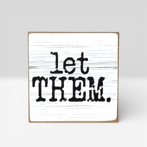 May include: A small, square wooden sign with the words "Let THEM." printed in bold, black letters. The background is white with horizontal wood grain lines. The sign has a natural wood-colored edge.