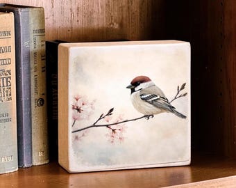 May include: A decorative wooden block featuring a detailed painting of a sparrow perched on a flowering branch. The bird has a brown head, gray and white body, and is set against a soft, neutral background. The block is displayed on a wooden shelf with vintage books.