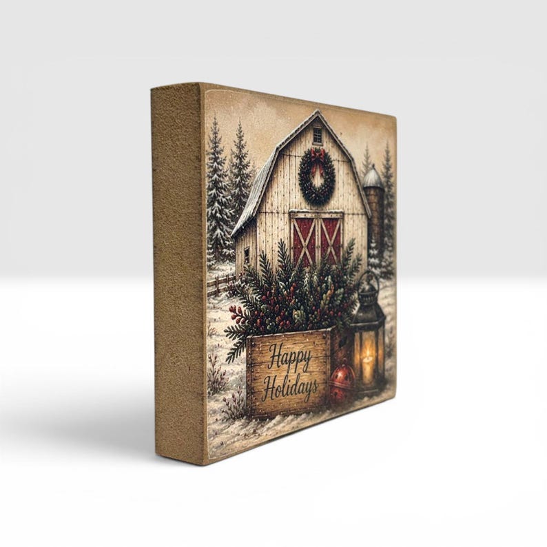 Puede incluir: Un letrero de bloque de madera rectangular con una escena invernal impresa de un granero blanco con una corona navide&ntilde;a, pinos y nieve. Una linterna y una caja de madera con el texto Happy Holidays est&aacute;n en primer plano.