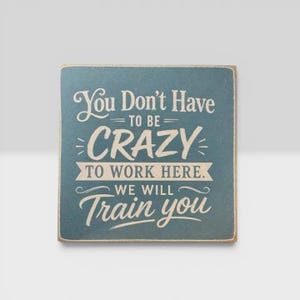 May include: A square, dark teal wooden sign with distressed edges featuring the white printed text: You Don't Have TO BE CRAZY TO WORK HERE. WE WILL Train you. The lettering uses a mix of serif and script fonts with decorative lines.