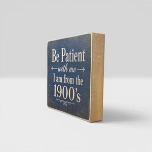 K&ouml;nnte beinhalten: Ein rechteckiges Holzblockschild mit dunkelblauer Vorderseite und naturfarbenen Holzseiten. Auf der Vorderseite steht in wei&szlig;er Schrift: Be Patient with me I am from the 1900's, verziert mit dekorativen Schn&ouml;rkeln &uuml;ber und unter dem Text.