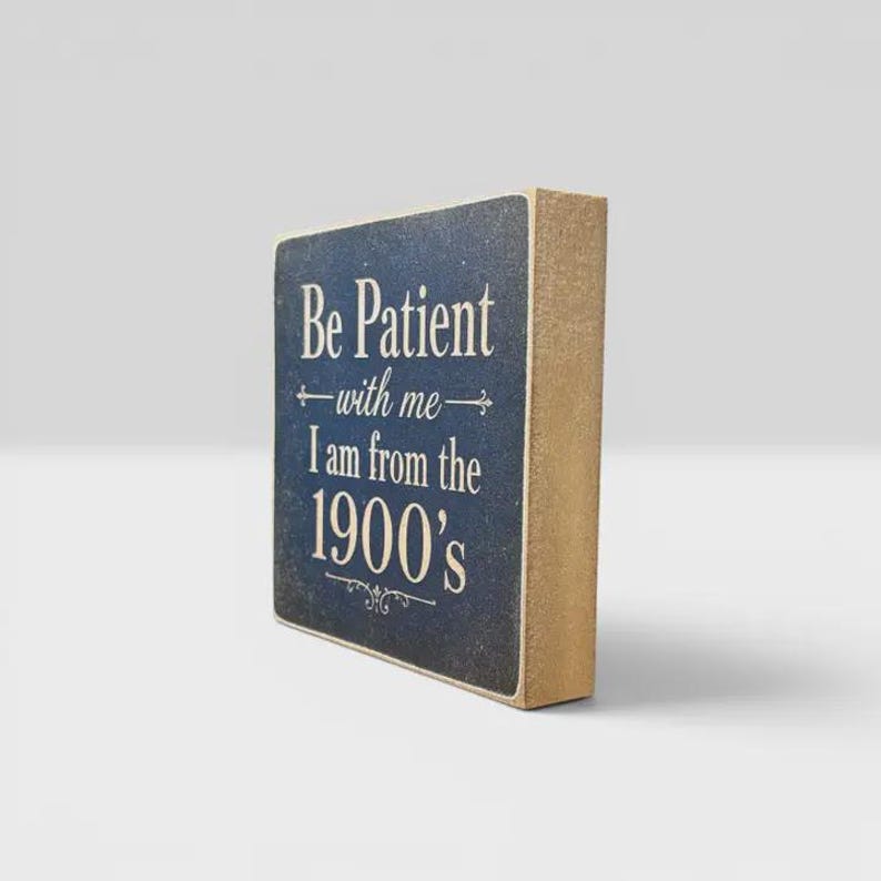 K&ouml;nnte beinhalten: Ein rechteckiges Holzblockschild mit dunkelblauer Vorderseite und naturfarbenen Holzseiten. Auf der Vorderseite steht in wei&szlig;er Schrift: Be Patient with me I am from the 1900's, verziert mit dekorativen Schn&ouml;rkeln &uuml;ber und unter dem Text.