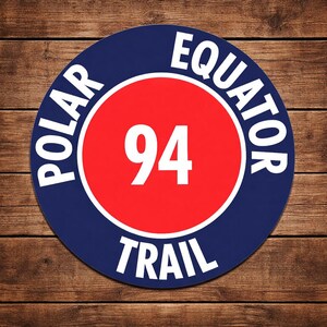 May include: A round sign with a blue border and white text that reads "POLAR EQUATOR TRAIL". Inside the blue border is a red circle with the number "94" in white. The sign is on a wooden surface.
