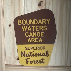 May include: A brown and yellow sign with the text "BOUNDARY WATERS CANOE AREA" and "SUPERIOR National Forest". The sign is mounted on a light-colored wooden surface.