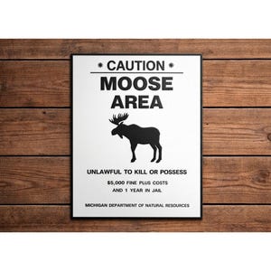 May include: A framed sign with a white background and black text that reads "CAUTION MOOSE AREA". Below the text is a silhouette of a moose. The sign also includes the text "UNLAWFUL TO KILL OR POSSESS" and "$5,000 FINE PLUS COSTS AND 1 YEAR IN JAIL".