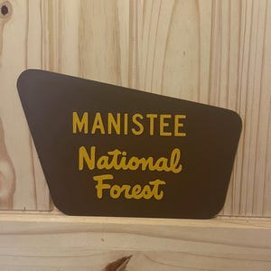 May include: A brown sign with the words "MANISTEE National Forest" in yellow lettering. The sign has a unique shape and is set against a wooden background.