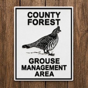 May include: A rectangular white sign with a black border featuring a black graphic illustration of a grouse in the center. The text reads COUNTY FOREST in the upper section and GROUSE MANAGEMENT AREA in the lower section in bold black lettering.