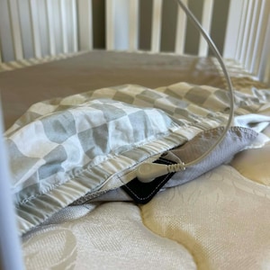 May include: A white crib with a beige mattress and a gray and white checkered blanket. A white cord with a connector is attached to the blanket. The crib has white vertical bars.