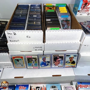 May include: Baseball trading cards in protective sleeves are displayed in cardboard boxes. The boxes are labeled with text including "Box 8", "R1", and "Baseball". Individual cards are visible, featuring players and team logos.
