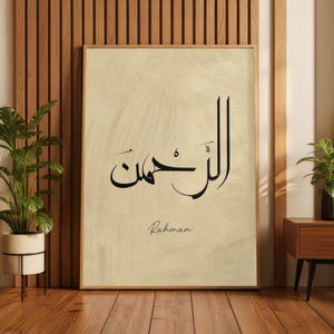 May include: Framed artwork featuring Arabic calligraphy in black ink on a beige background. The word "Rahman" is written in English script below the calligraphy. The artwork is displayed in a room with wooden paneling and a potted plant.