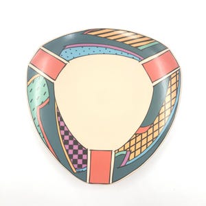 Puede incluir: Un plato decorativo triangular con un centro color crema y un borde verde azulado. El borde presenta patrones geométricos en naranja, morado, negro y verde. El plato tiene un diseño retro y abstracto.