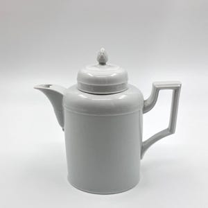 Può includere: Una teiera in ceramica grigio chiaro con coperchio e manico. La teiera ha un beccuccio e un piccolo elemento decorativo sulla parte superiore del coperchio. Il design è semplice e moderno, adatto per servire il tè.