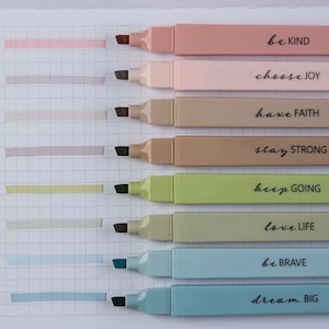 Puede incluir: Una colección de ocho resaltadores de colores pastel con frases inspiradoras impresas. Los colores incluyen rosa, melocotón, beige, marrón, verde, verde claro, azul y celeste. Cada resaltador tiene una frase correspondiente: "be KIND", "choose JOY", "have FAITH", "stay STRONG", "keep GOING", "love LIFE", "be BRAVE" y "dream BIG".