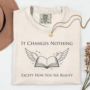 May include: Cream-coloured t-shirt with the text "IT CHANGES NOTHING EXCEPT HOW YOU SEE BEAUTY" and a graphic of a book with wings. A gold star pendant necklace and a brown hat are also visible.