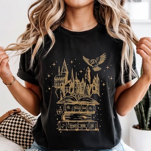 May include: Black t-shirt featuring a gold illustration of a castle, owl, and books. The books have the words "WIZARD BOOK" and "MAGICAL BOOK" printed on them. The shirt has a rolled sleeve design.