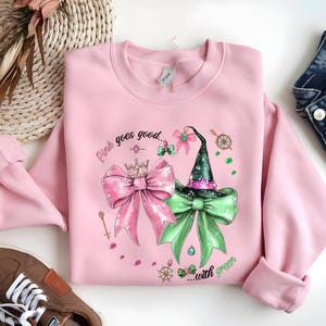 Peut inclure: Un sweat-shirt rose clair avec un motif graphique représentant un nœud rose avec une couronne, un chapeau de sorcière vert et un nœud vert. Le texte "Pink goes good...with green" est également présent.