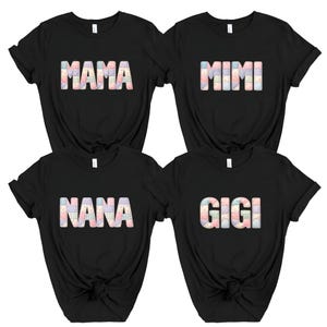 May include: Four black t-shirts, each displaying a different name: "MAMA", "MIMI", "NANA", and "GIGI". The names are rendered in a colourful patchwork style. The shirts are short-sleeved with a round neckline.